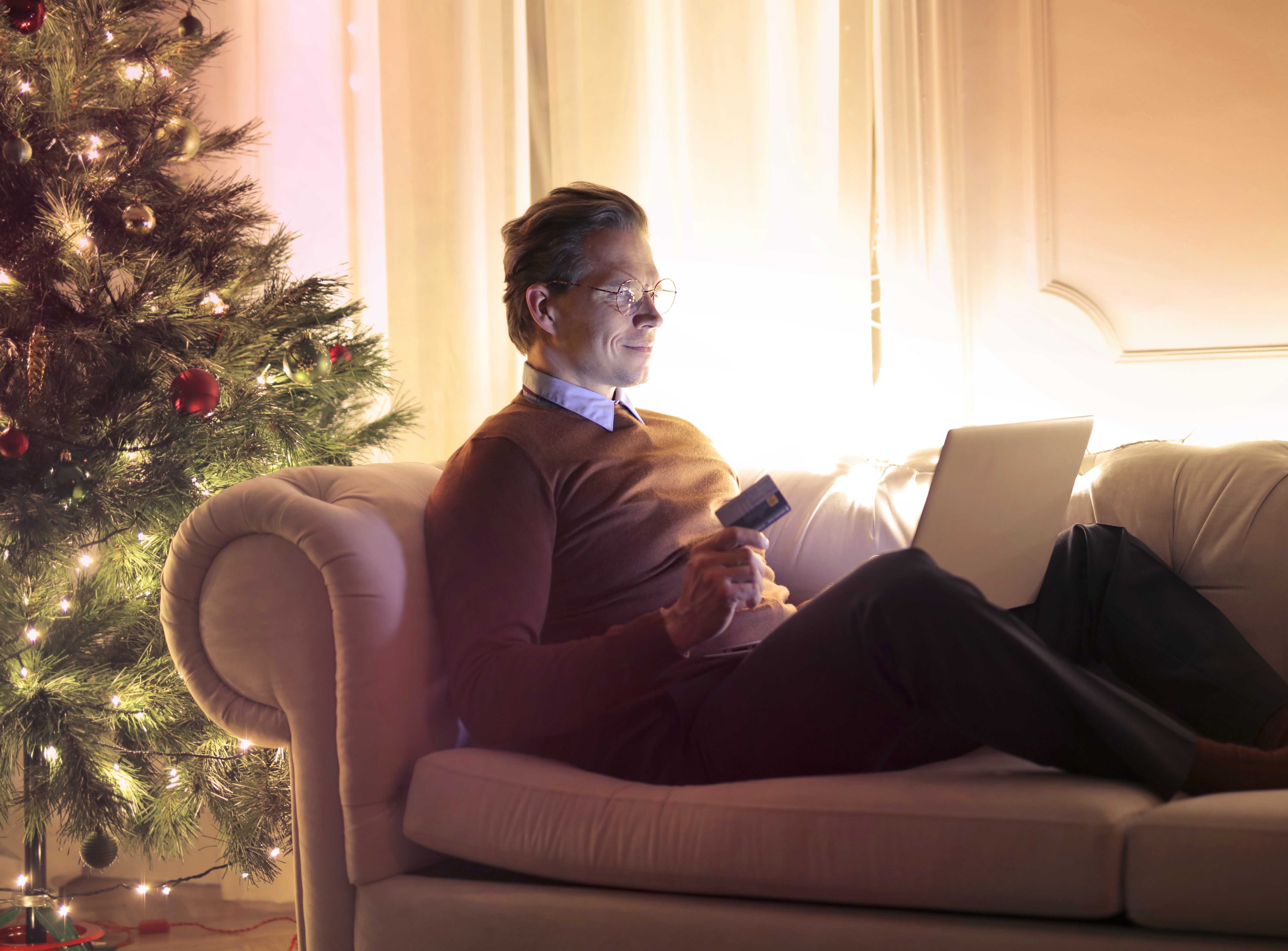 Man on Couch with Christmas Tree Lit in Background Holding Credit Card While on Laptop